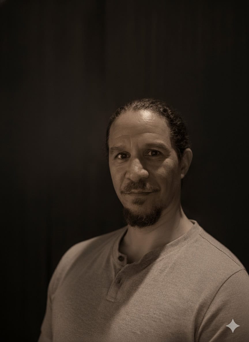 A man with a goatee and long hair pulled back, wearing a light-colored henley shirt, stands against a dark background.
